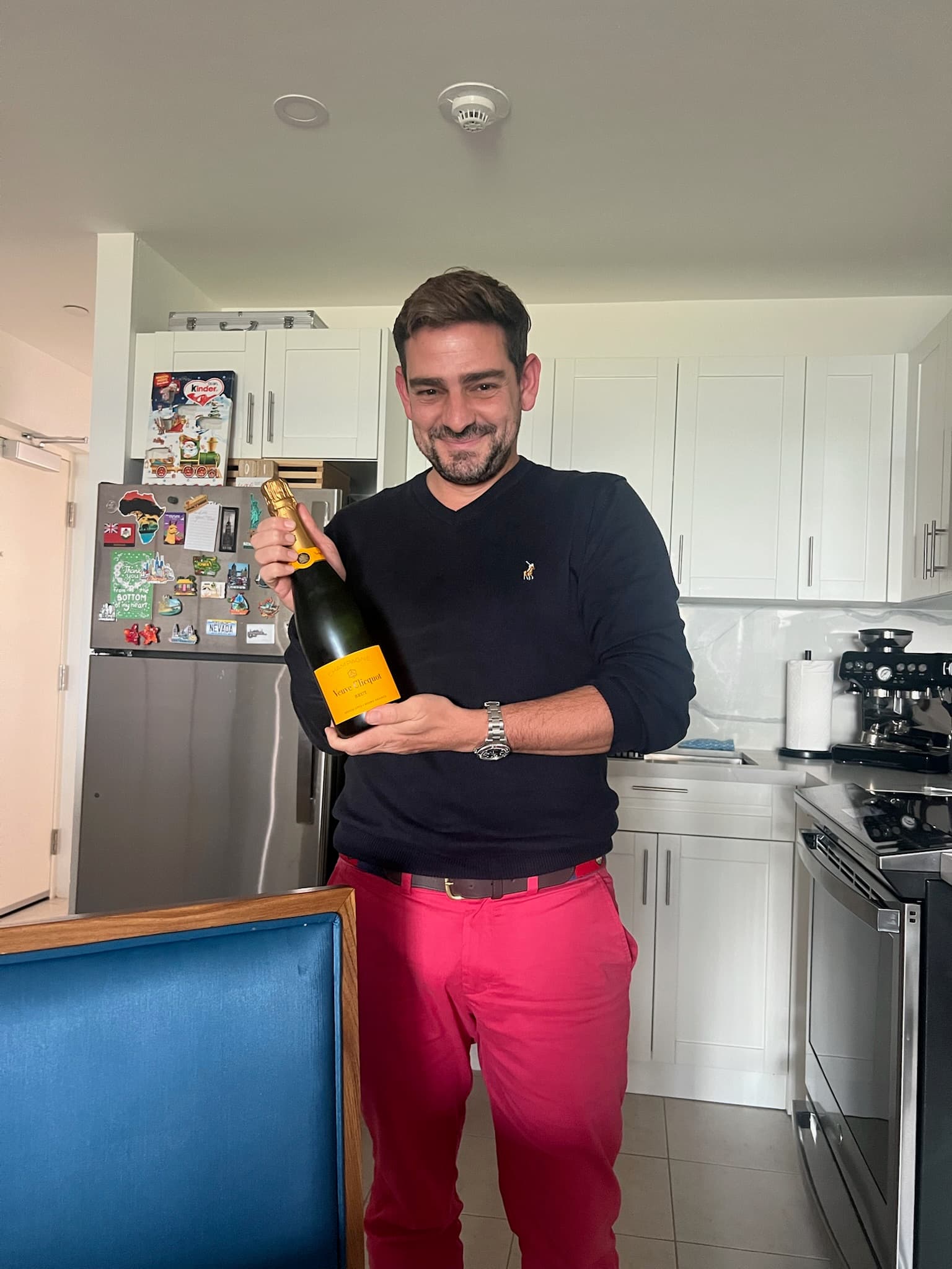 Smiling man in pink pants holding a bottle of Veuve Clicquot champagne in a kitchen.