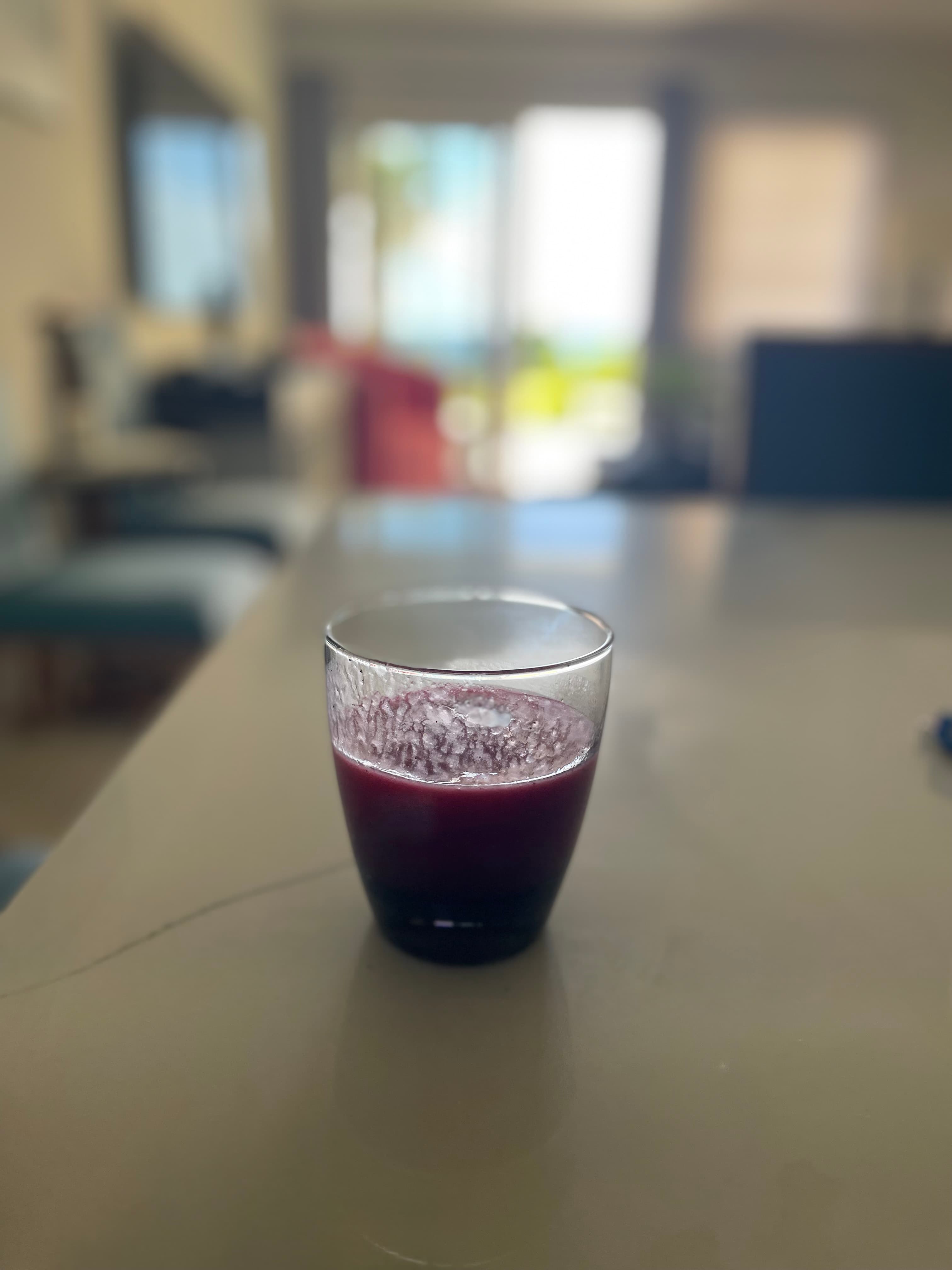 Dark purple juice in a clear glass on a light table with a blurred background.